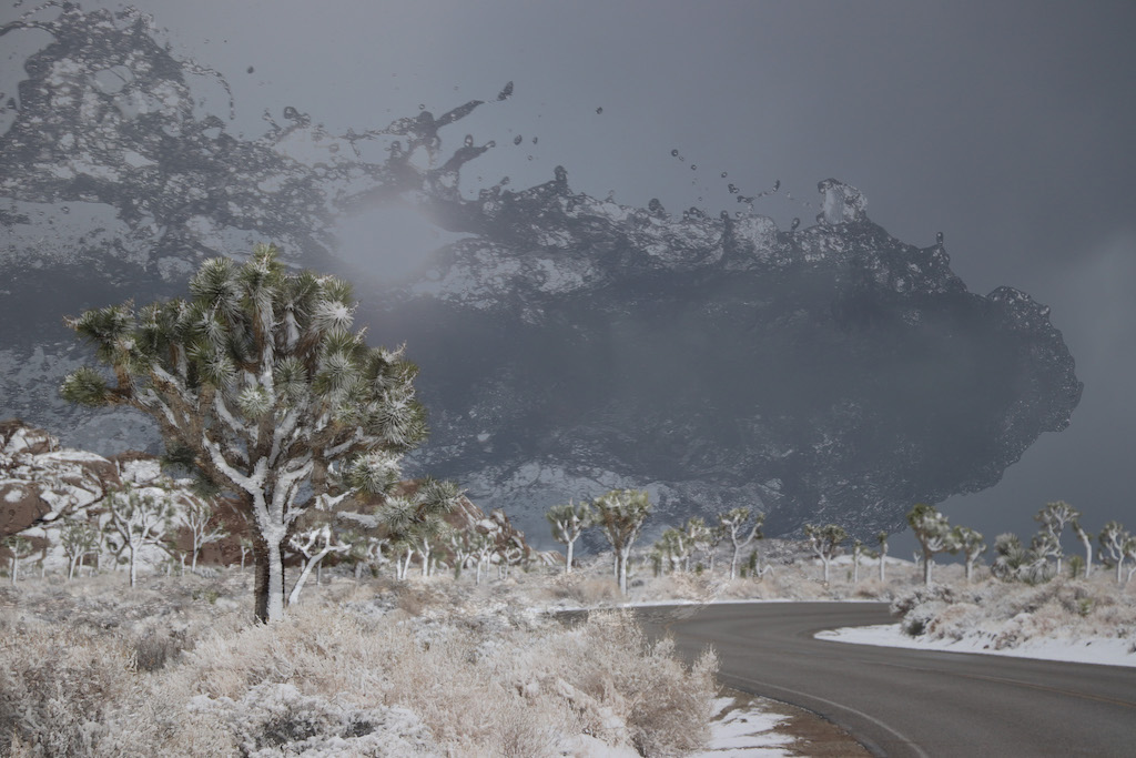 This image consists of two photos layered together.  The foreground is a scrubby landscape dotted by the occasional prickly desert tree.  In the bottom right, a highway winds away from the viewer to the horizon.  Snow dusts everything with white.  In the top half of the photo, where ordinarily clouds would be found, a larger-than-life translucent wave of water splashes across the grey sky. 