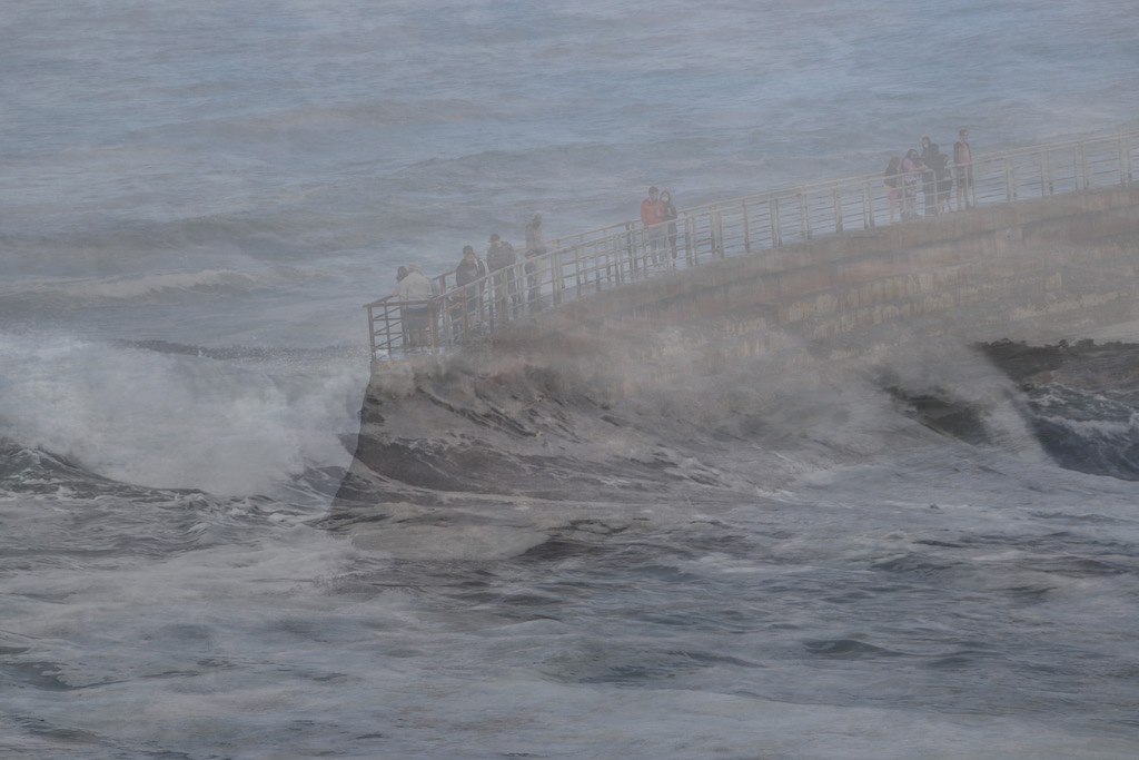 In monochromatic greys, two images are layered together.  Rising from the centre to the top right is a narrow but strong cement wall-like structure, possibly a dam, topped by a narrow walkway with railings.  Tourists stand against the railings looking down at their surroundings.  Superimposed on this scene is a rough translucent ocean-scape, so that it appears that powerful whitecapped waves are battering the structure and drenching the visitors with cold sea-spray.