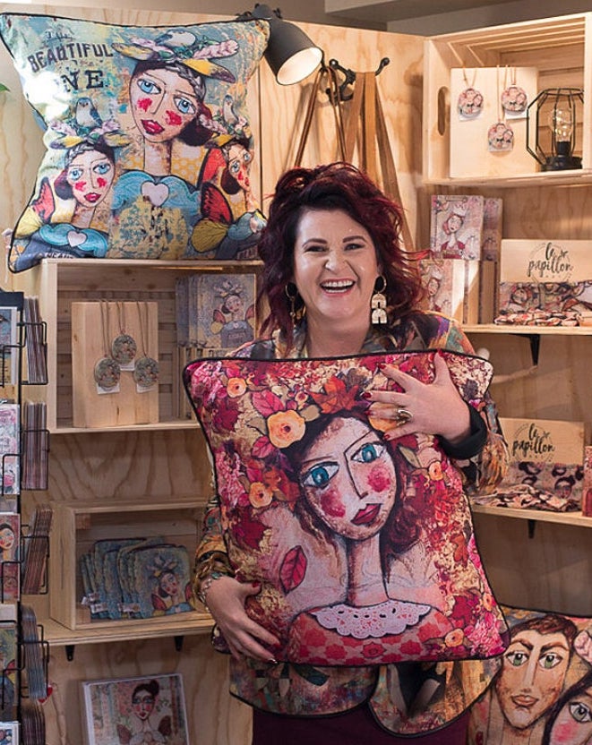 Photograph of Charlene du Toit in front of a store display of her artwork. Charlene is viewed from the knees up. She is wearing a multi-coloured blazer and is holding a decorative pillow with an image of her artwork printed on it. Charlene has red and black hair, is a white woman, is wearing large u shaped earrings and has a large smiles.