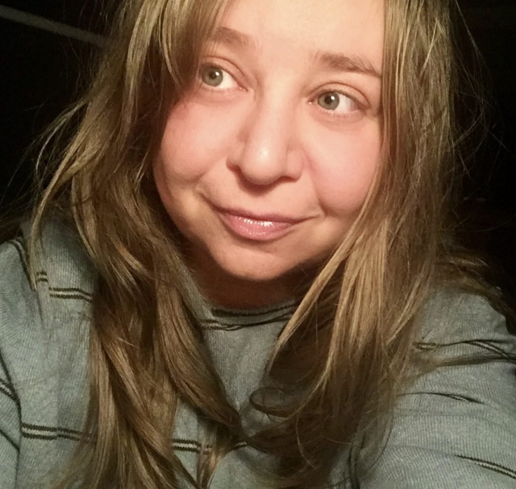 This is a photo of Sacha Kopelow.  She is a white woman, with long dark blonde hair, wearing a grey sweater.  She is glancing up to the side and smiling.