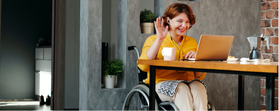 A woman in a wheel chair sits by a table with her laptop open waving and smiling at someone on the screen. The woman has white skin, short wavy red hair and is wearing a bright yellow shirt with beige pants.