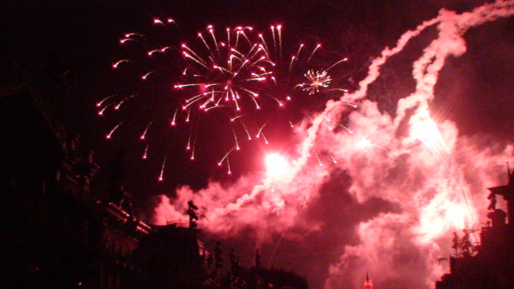 Photograph of red fireworks on a dark evening. There are clouds that may be smoke from the fireworks on either side of the image dark buildings can be seen from the reflection of the red fireworks.