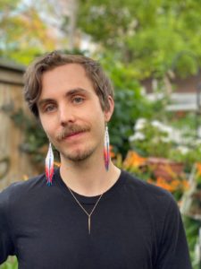 headshot of Bram Keast, Bram is a Caucasian male with short brown hair and a brown moustache. Bram is wearing long beaded earrings that are white and pink a well a a metal necklace. Bram is wearing a black long sleeved top and is standing outside with trees and bushes behind him.