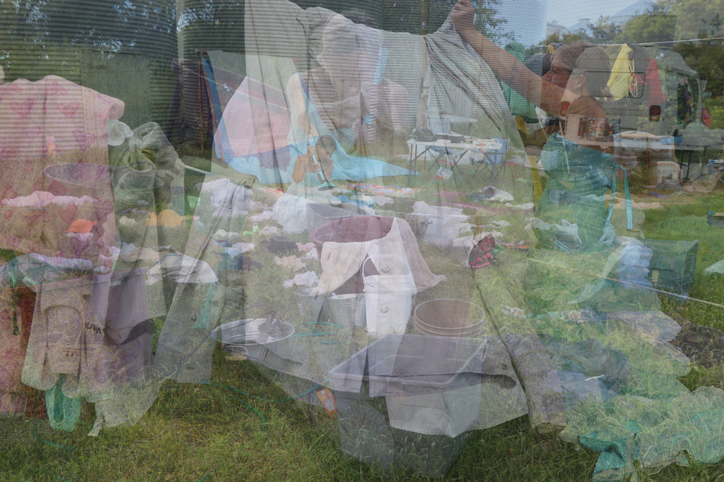 This image is made up of many photographs superimposed upon the other. Together photographs create a frantic image of Marie washing her laundry at different stages with laundry and buckets all over the area. Marie is washing her clothes in a grassy area with trees on the right-hand side and silos on the left-hand side. Marie is a thin white woman with long straight brown hair that is tied up. She is wearing a fancy blue dress that has lots of frills on the bottom. Marie can be seen using a blue plunger to wash her clothes and also seen hanging up her laundry to dry.