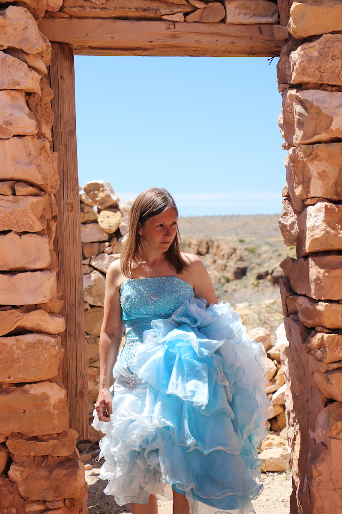 Photograph of Marie LeBlanc standing in the doorway of a rocky building. Marie is a thin white woman with long straight brown hair. Marie wearing a strapless blue dress with lots of frills at the bottom. The viewer is looking through the door to the bright blue sky and rocky terrain behind Marie.