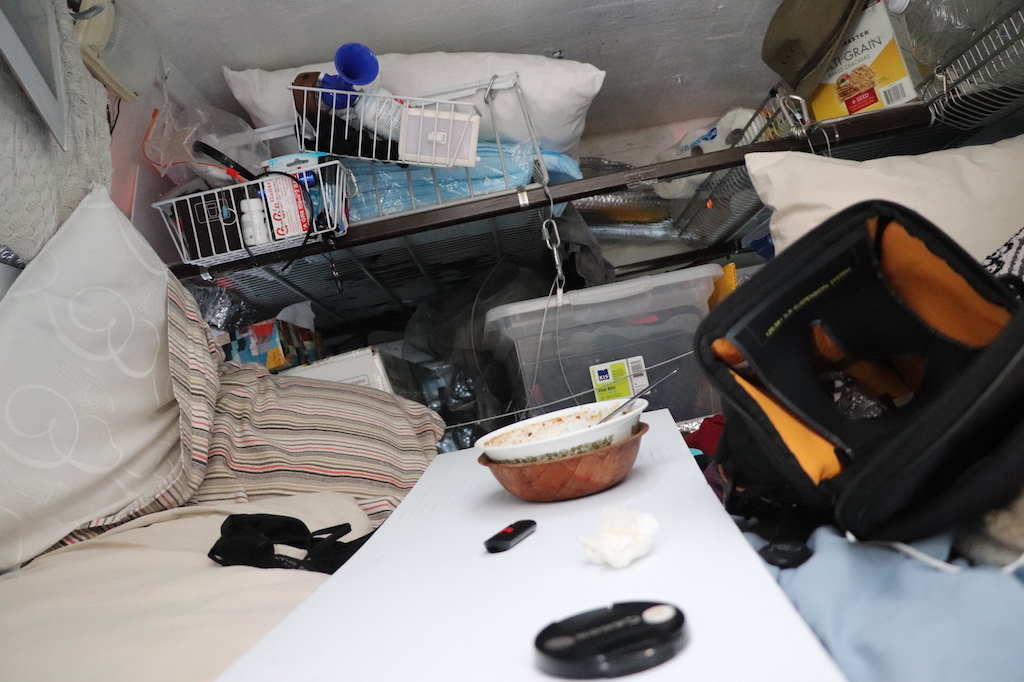 Photograph of the interior of Marie LeBlanc’s van. Her bed is visible and behind it are metal shelves with plastic bins full of supplies such as a blow horn, crackers, Kleenex, toilet paper and extra pillows.