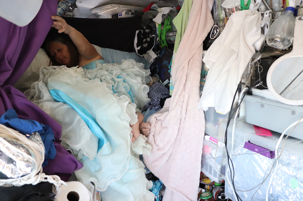 Photograph of Marie LeBlanc sleeping in her van. The van is packed with all of Marie’s possessions such as clothing, food, toilet paper and such. Marie is resting on her small bed that can just be seen behind all of the clutter. Marie is a thin white woman with long straight brown hair that is pulled back. Marie is wearing a blue strapless dress with lots of frills on the bottom.