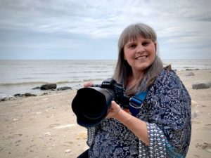 Headshot of Cheryle Broszeit. Cheryle is a white woman with long blond grey hair with bangs. Cheryl is at the beach and wearing a loose blue and white blouse and is holding a large Nikon camera. Cheryl has a large smile. 