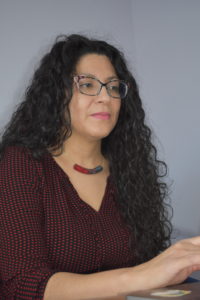 Headshot of Adriana Alarcón. Adrian is of Guatemalan descent and has long curly black hair with light brown skin. Adriana is wearing dark framed glasses with flowers, a beaded neckless and a black and red blouse 