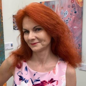 Headshot of Margaret Switala. Margaret us a white woman with red hair. She is smiling in the photo and is wearing a light pink top with flowers. Behind her I s white wall with artwork.