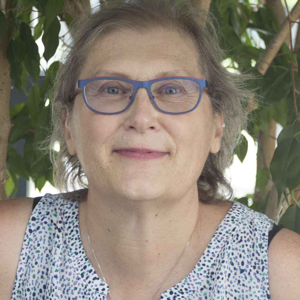 Headshot of Alice Crawford. Alice is a white woman with thin grey hair. She is wearing blue rimmed glasses and a dotted shirt. she is smiling and there are trees behind her