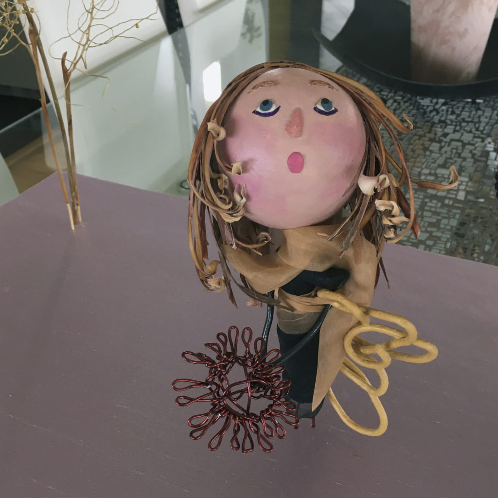 Photograph of a doll sculpture. The doll has white skin, blue eyes, blond/brown hair and a long tan scarf. She is wearing a long black dress. The doll is holding a flower that is created by twisting reddish coated aluminum wire.