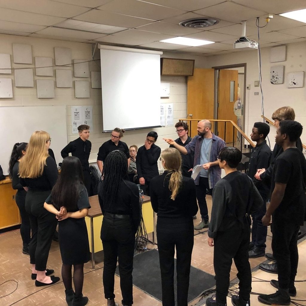 Photograph of a circle of people standing in a classroom. The choral members are all wearing black and the conductor is wearing casual clothing