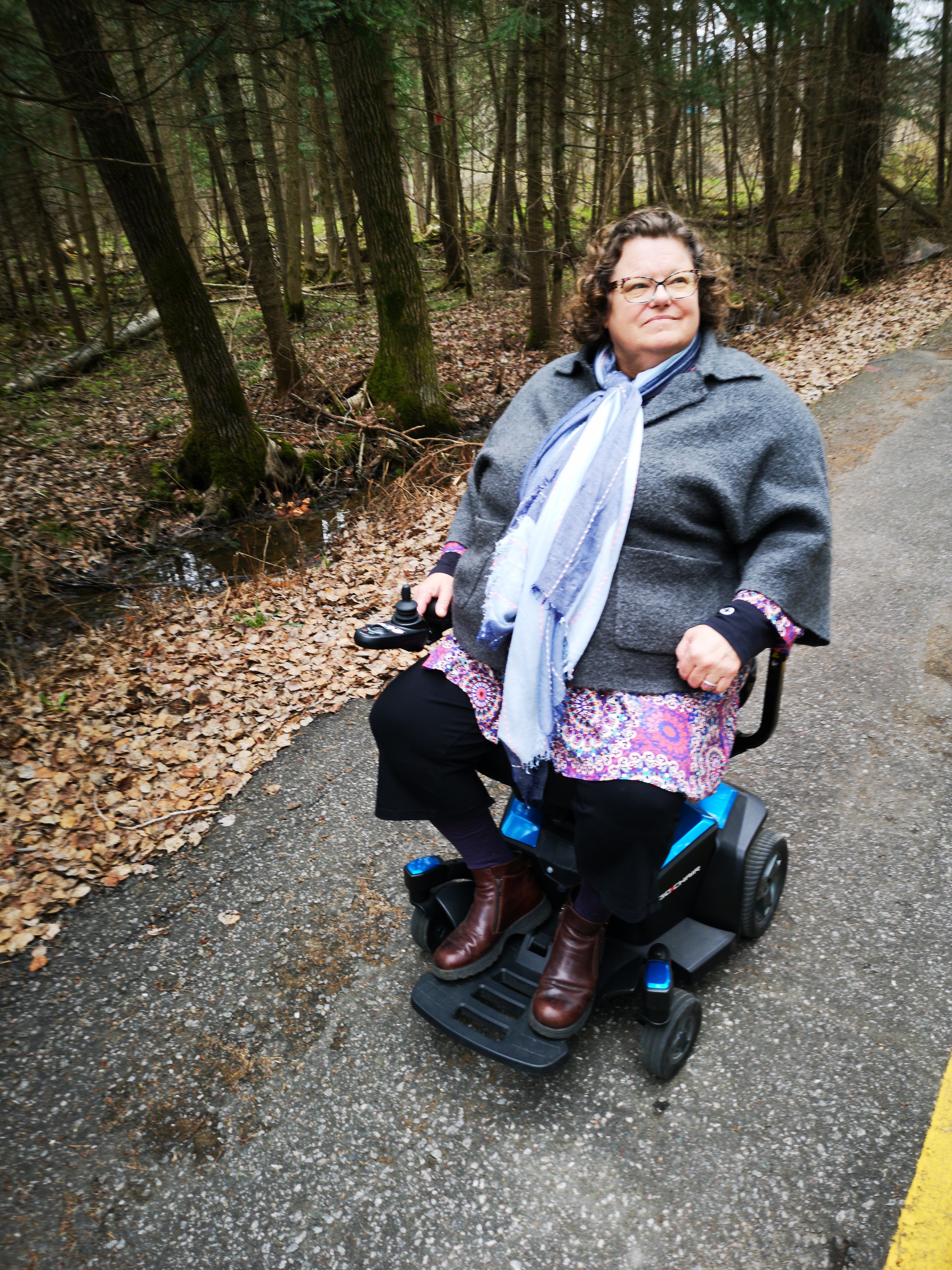 Photograph of Kim Kitchen on a path in a forest.  Kim is a white woman with medium length curly brown hair. Kim is wearing grey sweater over a colourful shirt, black pants and brown boots. Kim uses a scooter that is blue and black