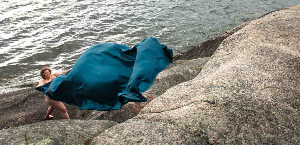 Photograph of Kim Kitchen from an upper perspective looking down on Kim. Kim is standing near a lake at the bottom of a rocky cliff. Kim is nude and is spreading out a large turquoise sheet that is billowing in the wind.