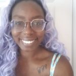 This is a photo of artist lita b. They are a Black femme, with long curly lavender-coloured hair. lita has a big friendly smile and is wearing glasses. They have a septum piercing and small tattoos on their collarbone and breastbone.