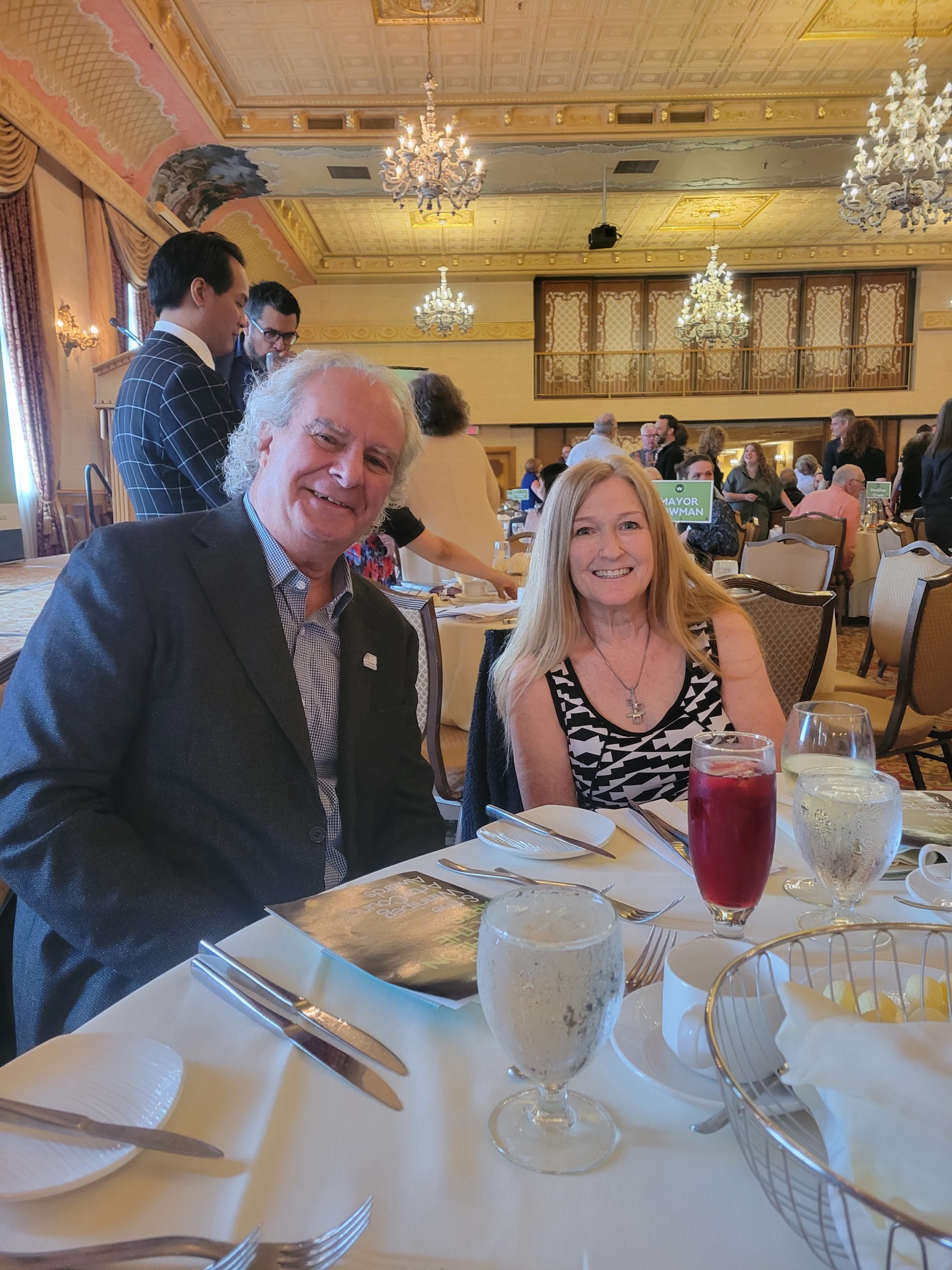 Photograph of Susan Lamberd and Robert Fabro at a dining table at the Fort Garry hotel. Robert is a white man in his 60s with grey hair and is wearing a grey suit jacket and stripped blue and white shirt. Susan is a white woman in her 50s with long strawberry coloured hair and is wearing a black and white dress. They are both smiling at the camera. In the background you can see many tables and other folks who attended the event. The space is very fancy with chandeliers, heavy curtains and a goldish ceiling.
