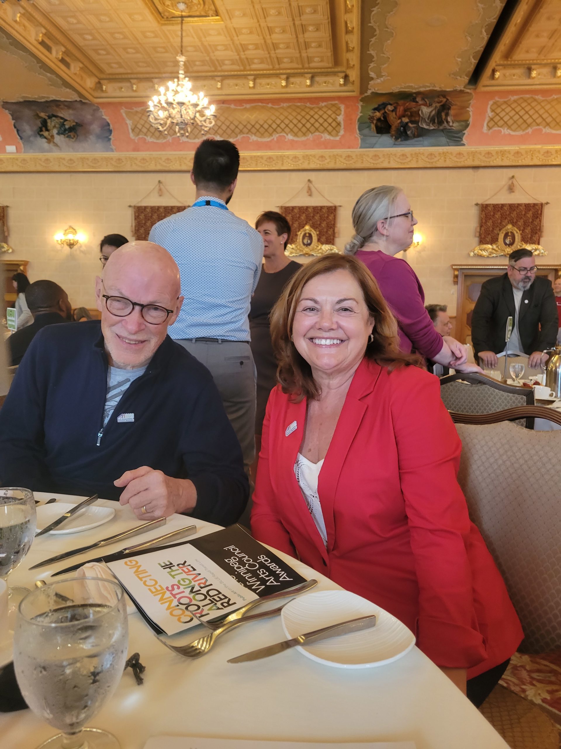 this is a photograph of Ted Howorth and Marie Bouchard. Ted is a white man in his 60s who is bald and black framed glasses. He is wearing a blue zip up sweater. Marie is a white woman in her 50s with shoulder length light brown hair. She is wearing a red blazer over a white t-shirt. They are both smiling at the camera. In the background you can see many tables and other folks who attended the event. The space is very fancy with chandeliers and a goldish ceiling.