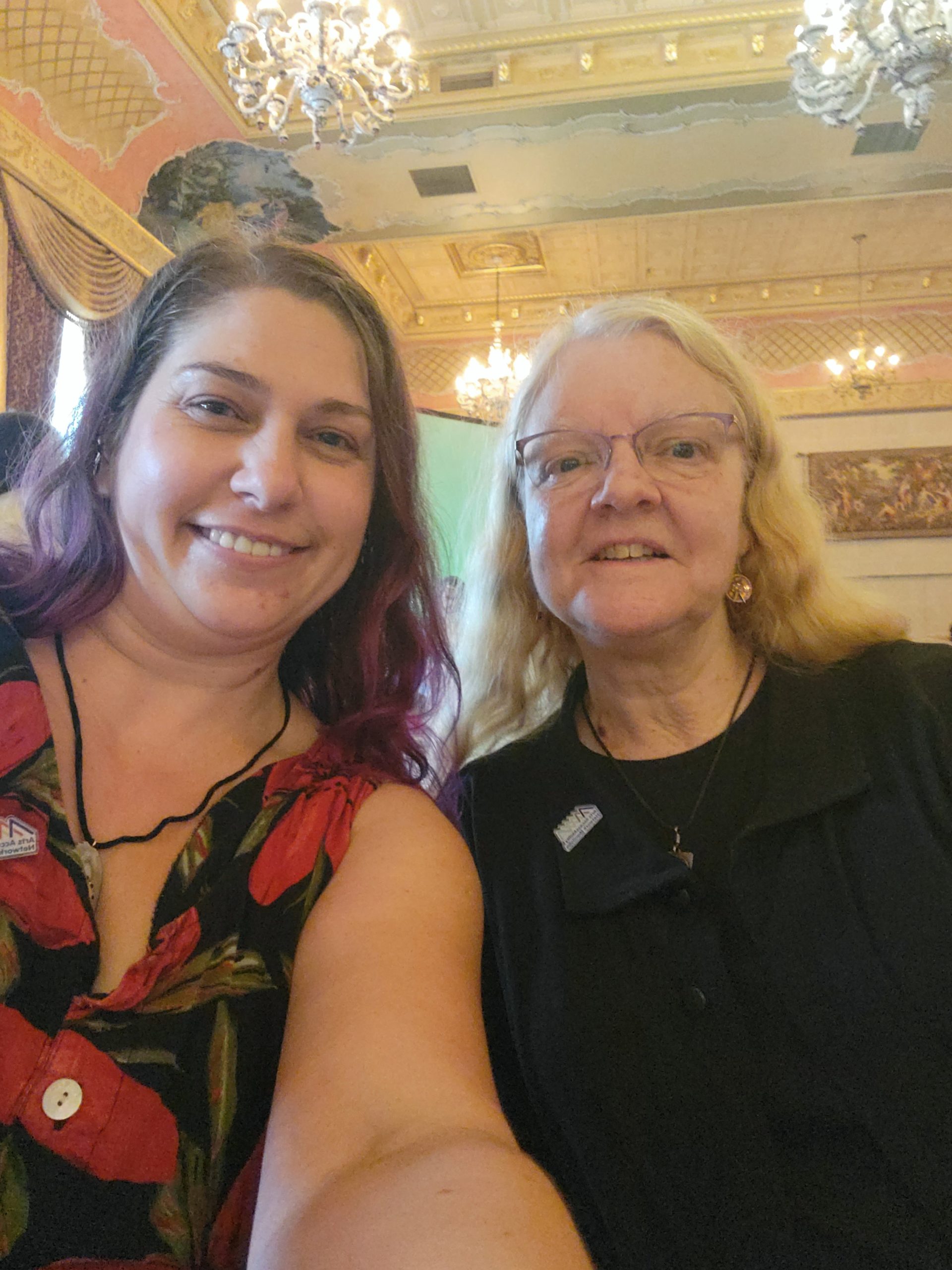 Photograph of Jenel Shaw and Diane Driedger. Jenel is a white woman in her 30s with long brown and purple hair. She is wearing a black dress with red flowers. Diane is a white woman in her 50s with long blond hair and purple rimmed glasses. She is wearing black sweater over a black shirt. They are both smiling at the camera. In the background you can see chandeliers and a goldish ceiling.