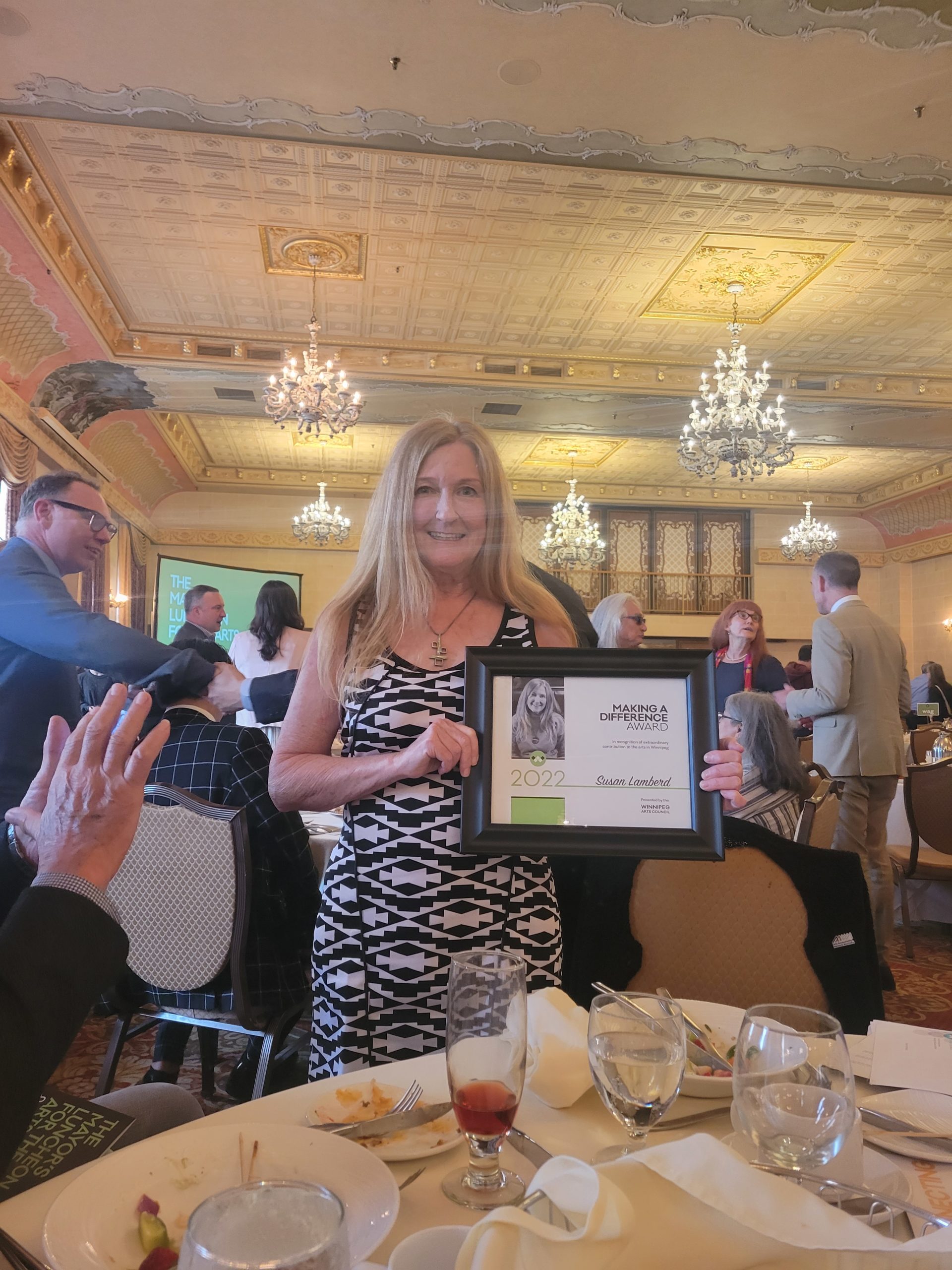 Photograph of Susan Lamberd holding her plaque award. Susan is a white woman in her 50s with long strawberry coloured hair and is wearing a black and white dress. They are both smiling at the camera. In the background you can see many tables and other folks who attended the event. The space is very fancy with chandeliers and a goldish ceiling.