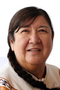 Head shot of Marie Annharte Baker. Marie is an Indigenous woman in her 60s with long brown hair that is braided and lays on her right shoulder. Marie is wearing a white shirt with symbols painted in black on the shoulders. Marie had brown eyes and is smiling at the camera 