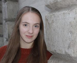This is a photo of dancer and choreographer Natalie Sluis. She is a young white woman. She is looking towards us with blue eyes and a shy smile. Her long straight light brown hair falls over her shoulders. She wears a simple dark orange sweater.