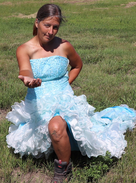 This is a photograph of Marie LeBlanc. Marie is a white woman with brown hair tied back. She is kneeling and holding out a hand invitingly to the viewer. She is wearing a fancy blue dress and work boots. 