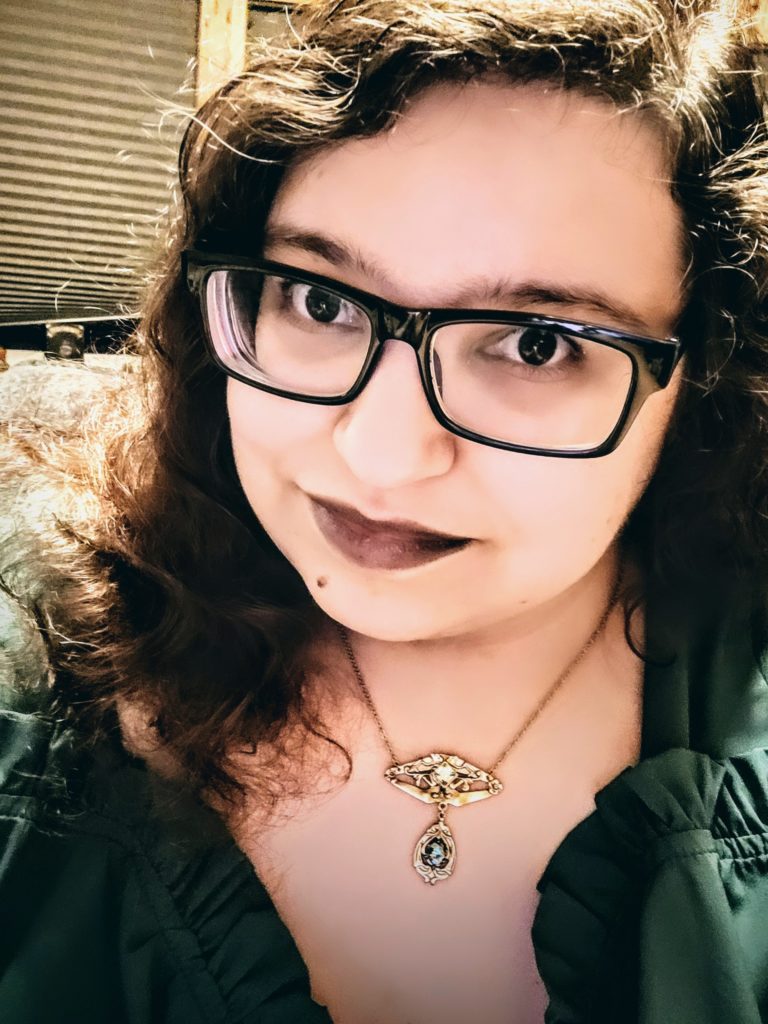 headshot of Meagan Hoskins who is a white woman with curly brown hair. She is wearing square black glasses, a dark green shirt and has dark purple lipstick.