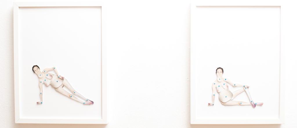 This is a photograph of two framed artworks. Each artwork is a paper doll of Yvette Cenerini. The paper dolls have been placed in different positions. The doll on the left shows Yvette on her side while pushing herself up with one hand and the other hand on her hip. The doll of the right is sitting down with one leg stretched out and the other bent with her hand on her knee.