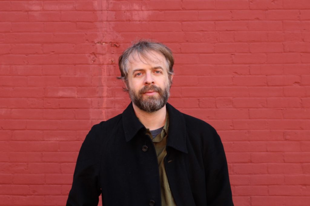 Photograph of Jacob Scheier. Jacob is a white man in his 50s with brown hair and a beard with streaks of grey. Jacob is wearing an olive-green shirt under a black button up top. Jacob is gazing into the camera with a slight smile. He is standing in front of a brick wall painted red.