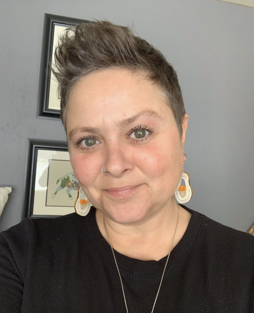 Headshot of Candace Lipischak. Candace is an indigenous woman in her 50s with short brown hair mixed with grey. She has bright grey/green eyes and is smiling at the camera. She is wearing bone earrings with orange beads and a black shirt.