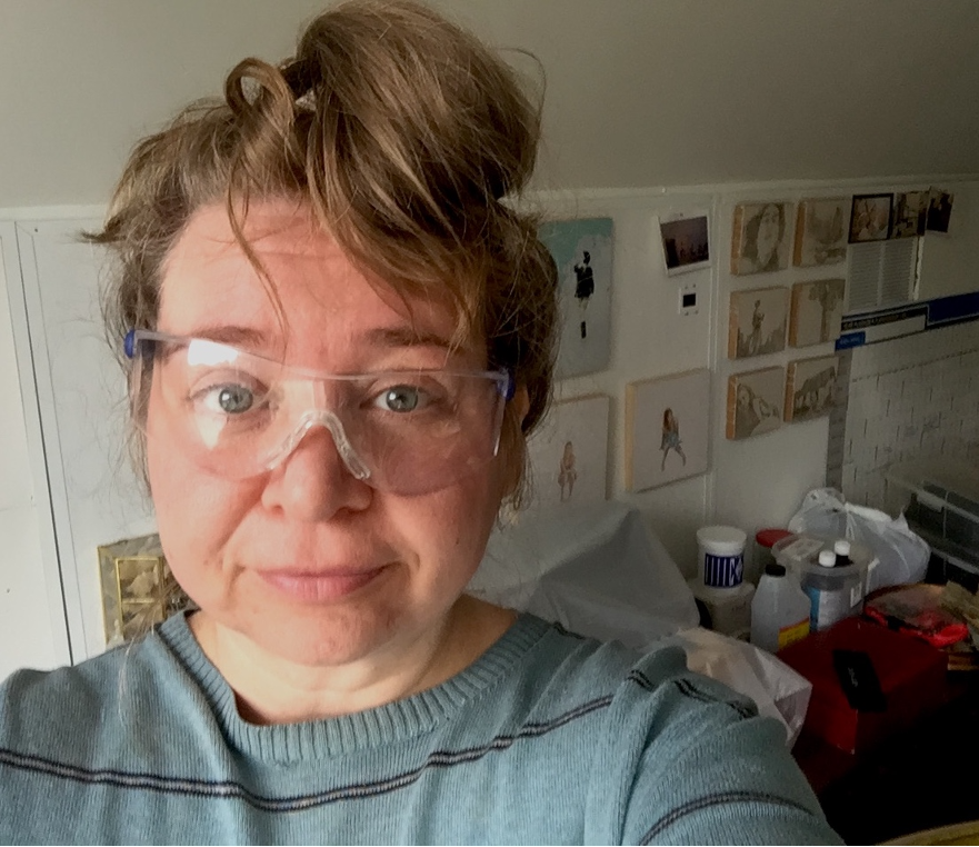 This is a selfie headshot of artist Sacha Kopelow in her studio. Sacha is a middle-aged white woman, with a shy smile. She wears safety goggles slightly askew, and her long dark blond hair is tied up in a messy bun, some strands falling down over her eyes. Behind her we glimpse some of her drawings and paintings on the white studio wall, a yearly-planner calendar, and a jumble of art supplies.