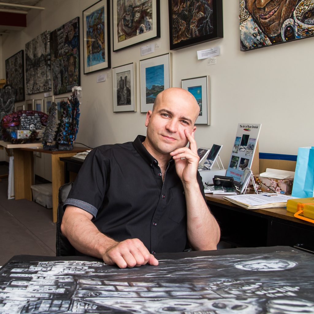 Photograph of Ryan Smoluk in this art studio. Ryan is a white male in his 30s. He is bald and is wearing a black button up short. Ryan is sitting at his desk with his colourful artwork behind him.