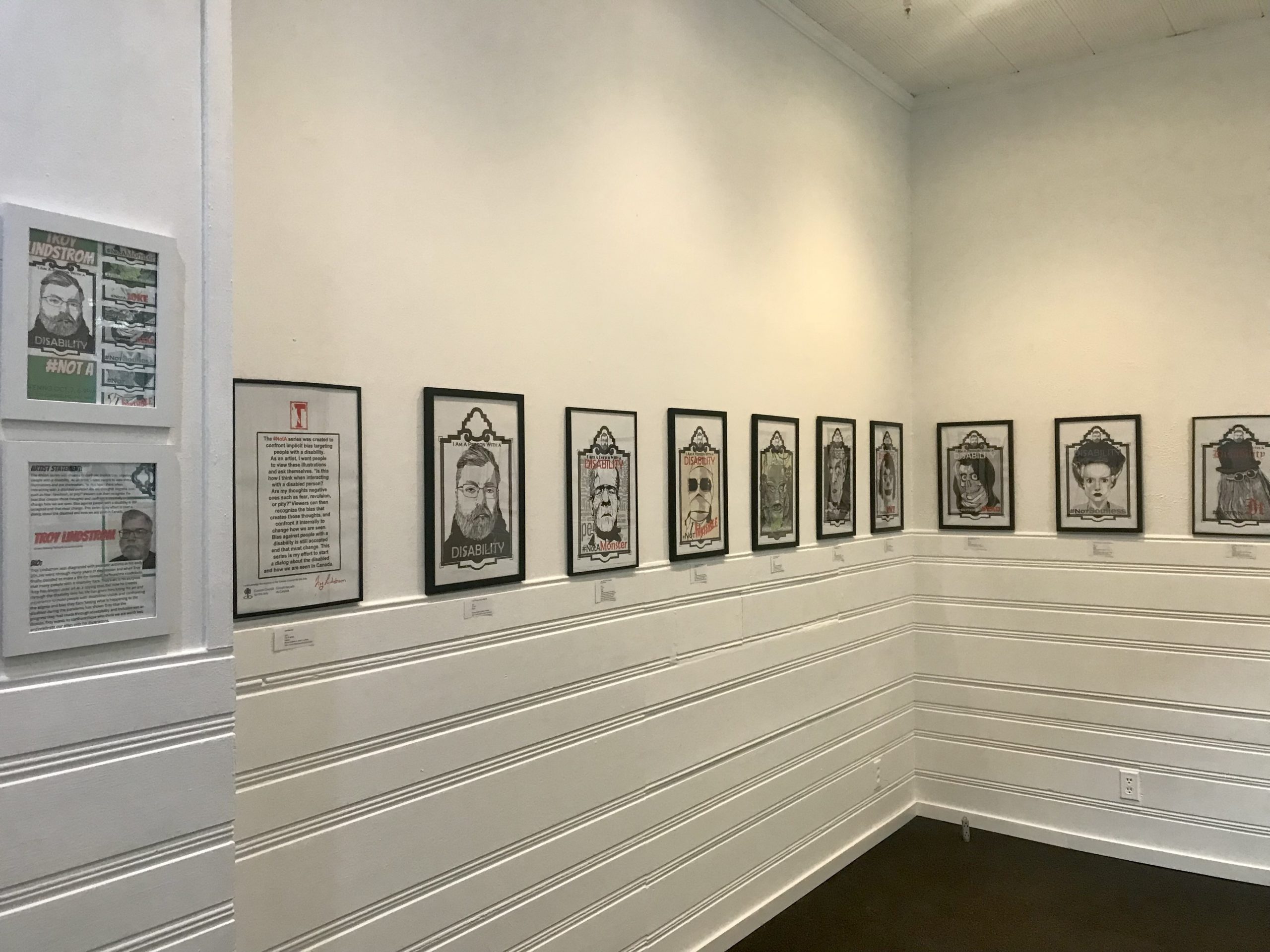 This is a photo of the AANM Gallery's white walls, displaying the graphic artwork of Troy Lindstrom in black frames.