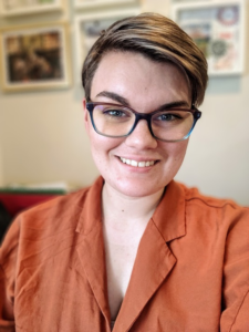 This is a headshot of Jen Sebring (they/them). Jen is white and has short brown hair. Jen is wearing black rimmed glasses and is smiling the camera