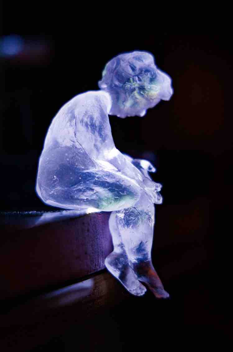 A fat translucent cast glass woman sits on a sheer edge, legs dangling off into darkness. Her head is bowed and her hands are in her lap, empty palms up.