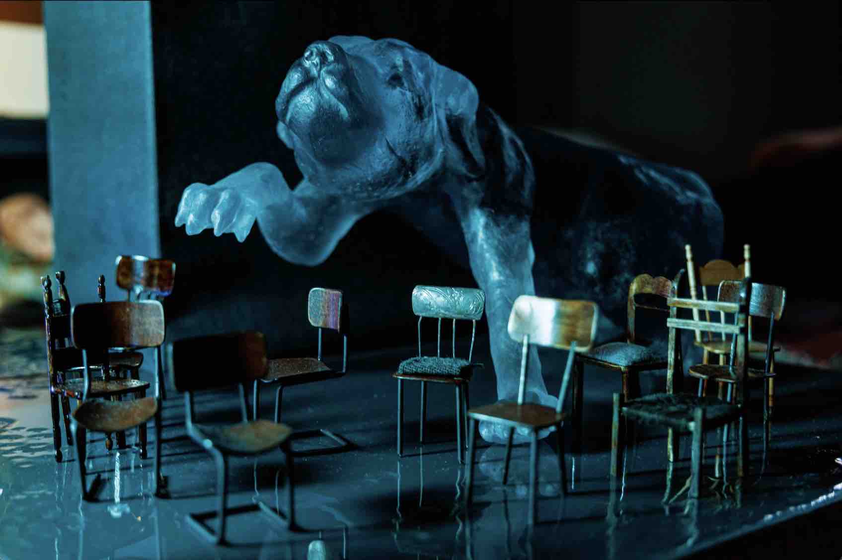 A large blue cast glass dog reaches a paw tentatively over a theatre of small empty chairs. Each chair is about the size of his paw, and woodworked in a different style. The lighting is eery, dim and green-blue, causing the chairs to cast interesting shadows.