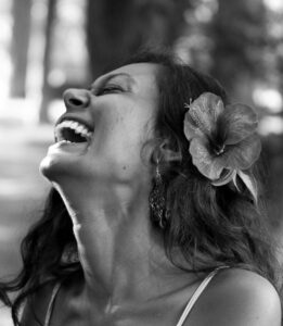 Photograph of Shayani Turko. Shayani is a East Indian woman with long dark hair. The image is of Shayani's profile while she is laughing. her hair is loose and has a large flower being her ear in her hair.