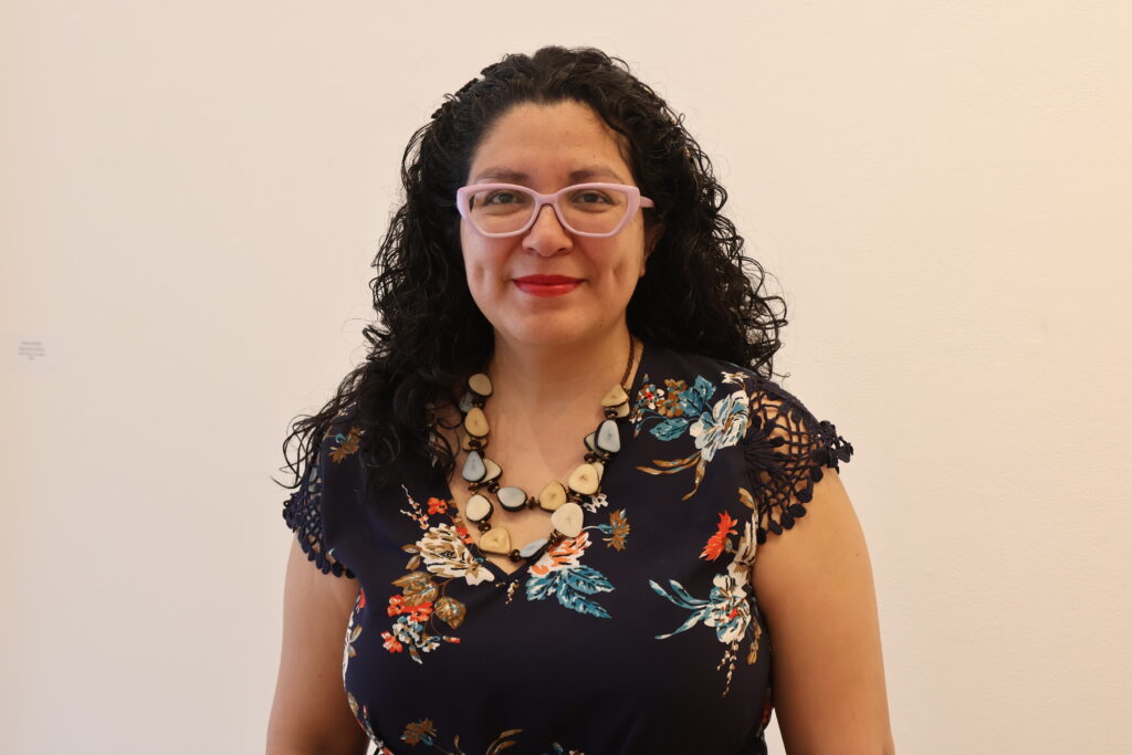 This is a photograph of Adriana Alarcón. Adriana is a Guatemalan woman in her late 30s with beautiful curly black hair and clear caramel skin. Adriana is smiling and her dimples are shining through. She is wearing pink rimmed glasses, a dark shirt with a floral motif and a necklace with many stones.