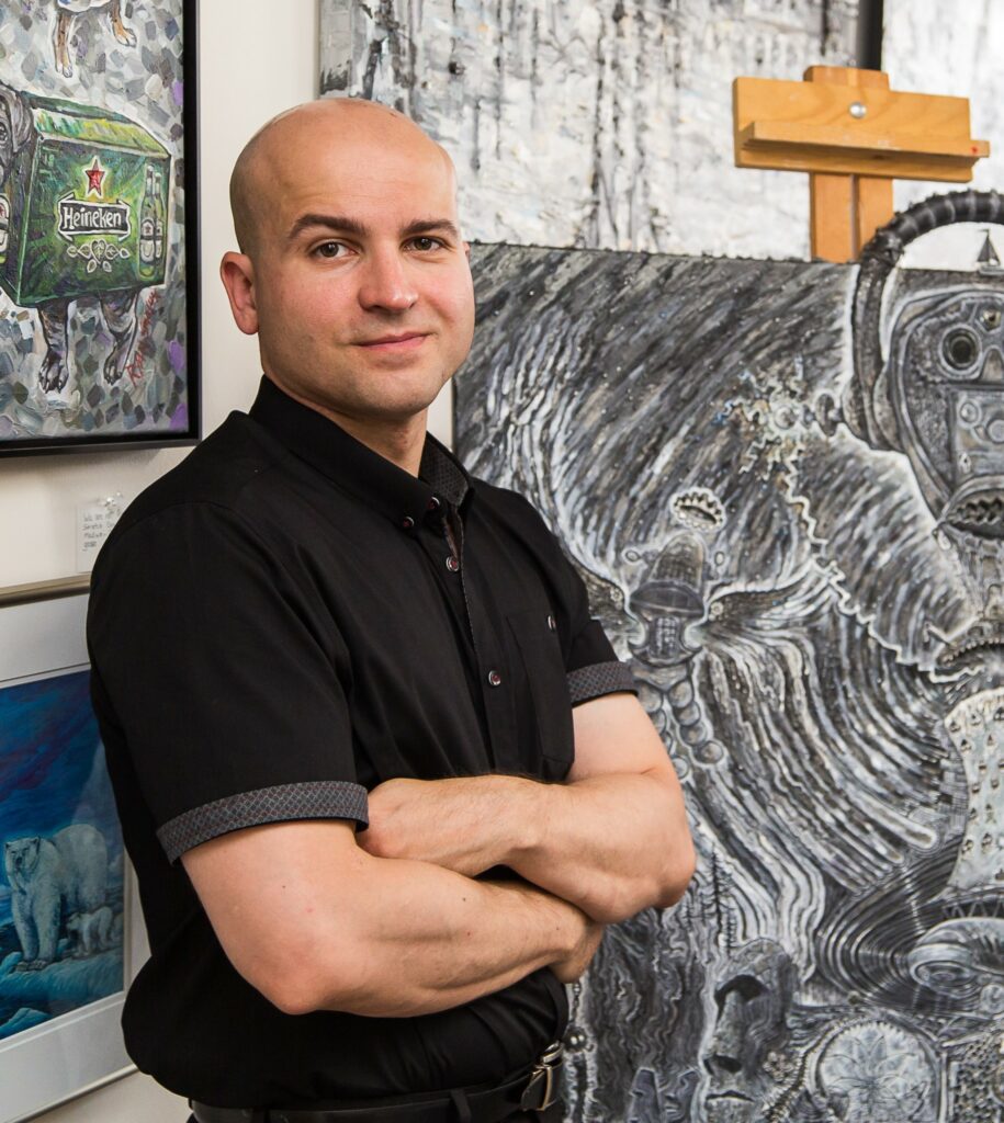 This is a photograph of Ryan Smoluk in his studio. Ryan is a white man in his 40s. He is smiling slightly at the camera and stands with his arms crossed. Ryan has a shaved head and clean shaved face. He is wearing a black button up top. Ryan is standing in front of his artwork which features many intricate details.
