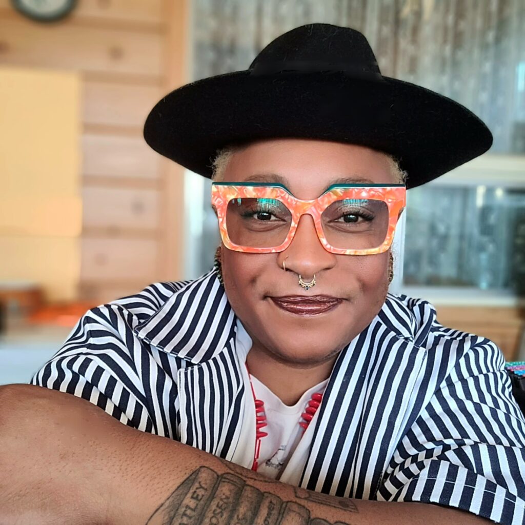 This is a photograph of m. patchwork monoceros. mel is a black woman in her 40s or 50s and is smiling and looking directly at the viewer. mel is wearing a black hat with a rim, funky orange glasses, a black and white striped shirt and jewelry and piercings on ger ears and nose.