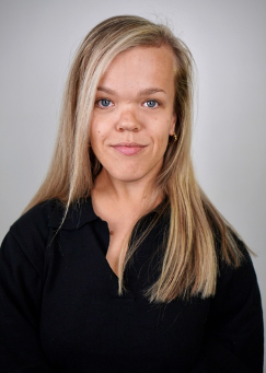 A headshot of Alexia, a white, Disabled woman with blonde hair. She is staring directly at the camera with a slight smirk. She's wearing a black shirt with gold hoop earrings. The background is white. Photograph by Lindsay Wu.