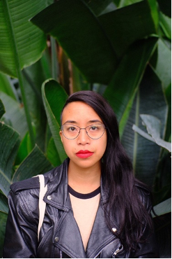 Headshot of Andi Vicente (they/them). They are a person of colour with long dark hair and bright red lipstick.  Andi is wearing wire-rim glasses and a black leather jacket, and standing before a background of green banana leaves.