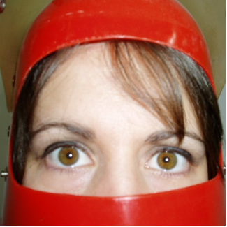 This is a photo of Erika-Jean, slightly out of focus and at close range. She is wearing a bright red plastic helmet, so that all we see of her face are her wide, serious hazel eyes and her forehead, partially covered by her brown bangs. She is a light-skinned woman.