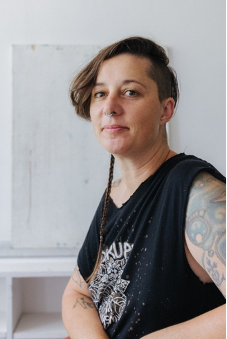 Headshot of Michelle Wilson. Michelle is a white woman with a punky haircut, long on one side and shaved above her left ear, with a small braid hanging down her shoulder. She is seated at an angle but looking directly into the camera with a friendly, bemused manner.  She has tattoos on her bare arms and is wearing a black cutoff t-shirt.