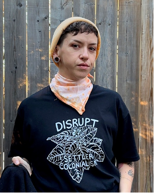Photo of Sheri Nault (they/them). Sheri is pictured casually in front of a weathered wood fence. Sheri is Métis and is wearing a black t-shirt which reads ‘DISRUPT SETTLER COLONIALISM’ in white lettering over a drawing of a tobacco plant. Sheri looks at us with a serious expression. They wear a nose ring, ear spacer, orange neckerchief, and orange cap. Tattoos are visible on their arm.