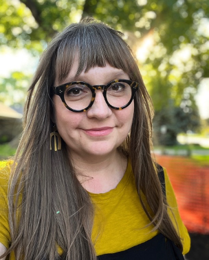 Headshot of Stacey Abramson.  She is a light-skinned woman with long straight light brown hair and heavy round tortoiseshell glasses.  She looks directly towards us with a friendly smile. Behind her are trees and dappled sunlight, out of focus.