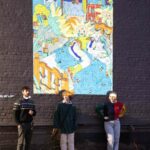 Three young people lean against the grey brick of the AANM Gallery building parking lot wall. Above them, a digital drawing by Jude Palace, colourful and chaotic, is projected onto the wall.