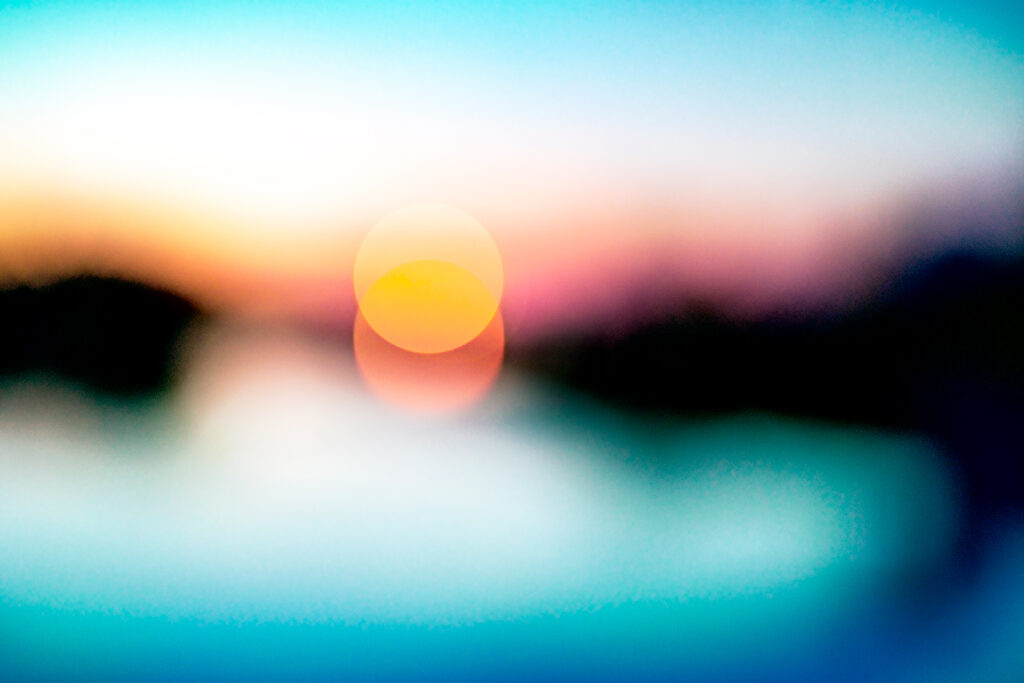 This photograph appears abstract because it is blurry, out of focus. In the centre, light has formed itself into two perfect circles of yellow and orange. On either side are fuzzy black forms, possibly a landmass. Below is a fuzzy blue expanse (a sea?), above is fuzzy pink, orange, white and aqua (sky?).