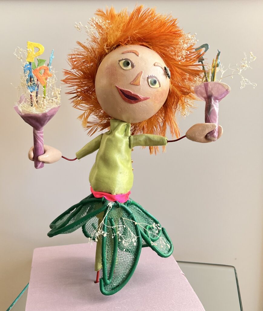 Feathered ginger hair in light green top with a neon pink sash at the waist. The dark green petal like skirt is covered with netting stretched over dark green wire spelling Gerlin. She wears light green capris under her skirt. She is of pale complexion with rosy cheeks, a longish nose with a snowflake stud, green eyes famed by brown eyelashes. Gerlin is happy with her mouth slightly open. In each hand, she is holding out a bouquet of flowers composed of brightly coloured letters spelling PARTY and baby’s breath.